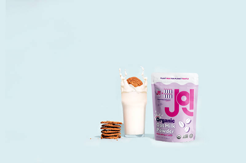 A glass of oat milk with a cookie splashing in it next to a JOI Organic Oat Milk Powder pouch.