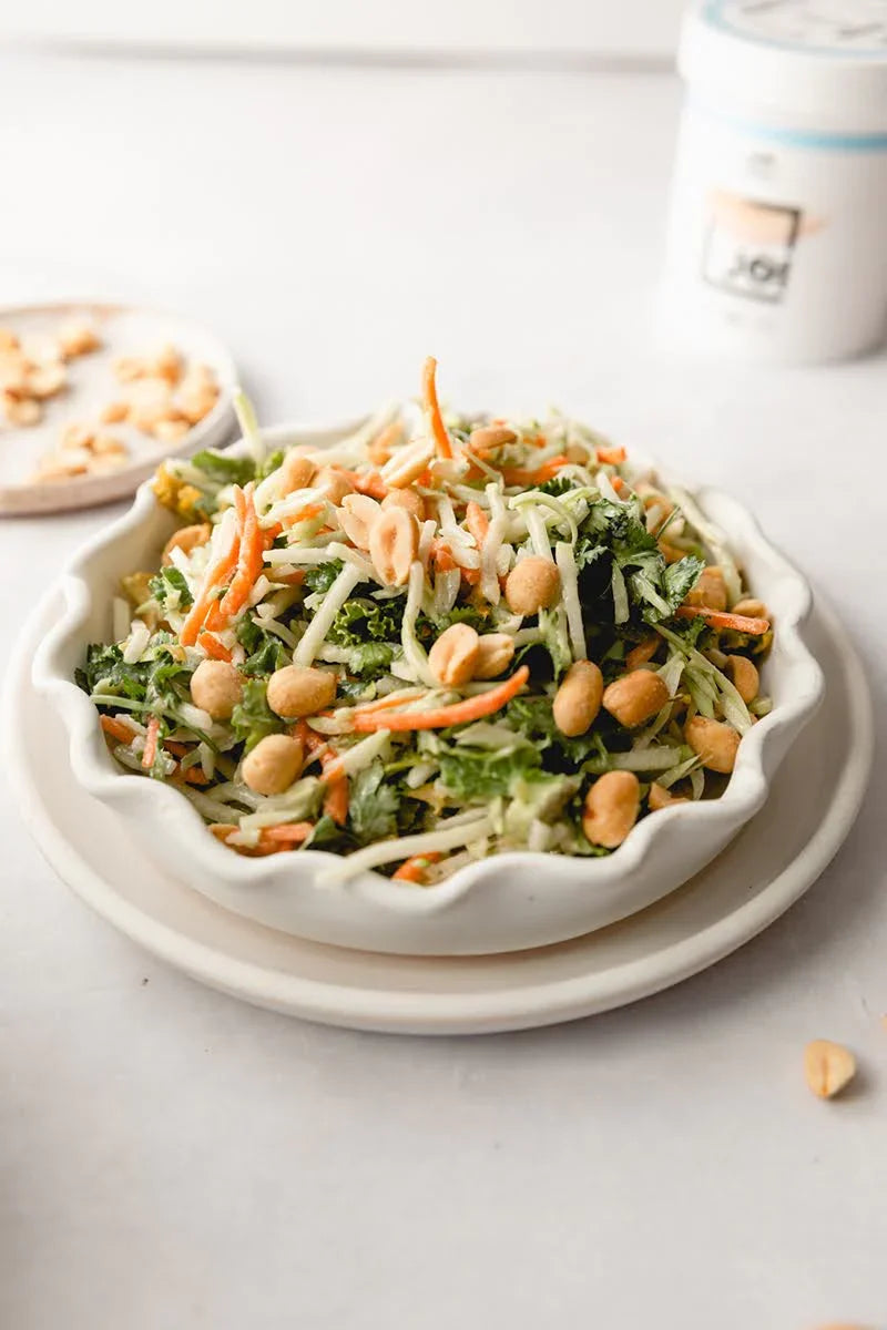 A white bowl of broccoli slaw with sesame ginger soy dressing, carrots, and peanuts, with a JOI carton.