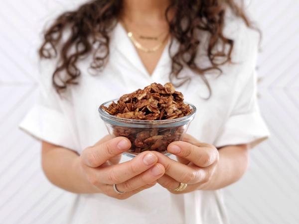 A woman holds a small bowl of crispy chocolate sea salt granola made with JOI, against a white background.