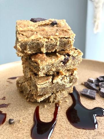 A stack of Gluten Free & Vegan Date Sweetened Blondies, drizzled with chocolate sauce on a tan surface.
