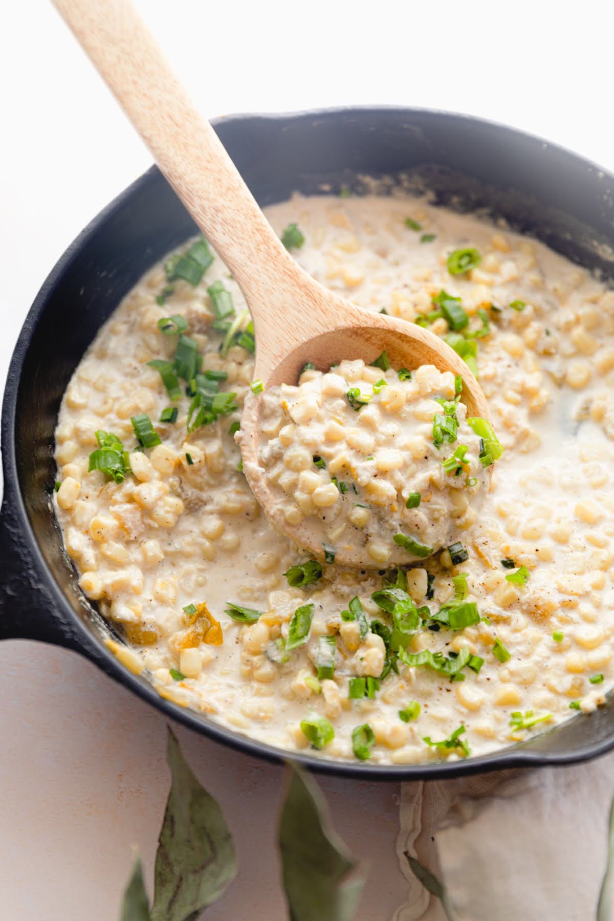 A wooden spoon lifts creamy JOI dairy-free creamed corn with green chiles and chives from a black skillet.