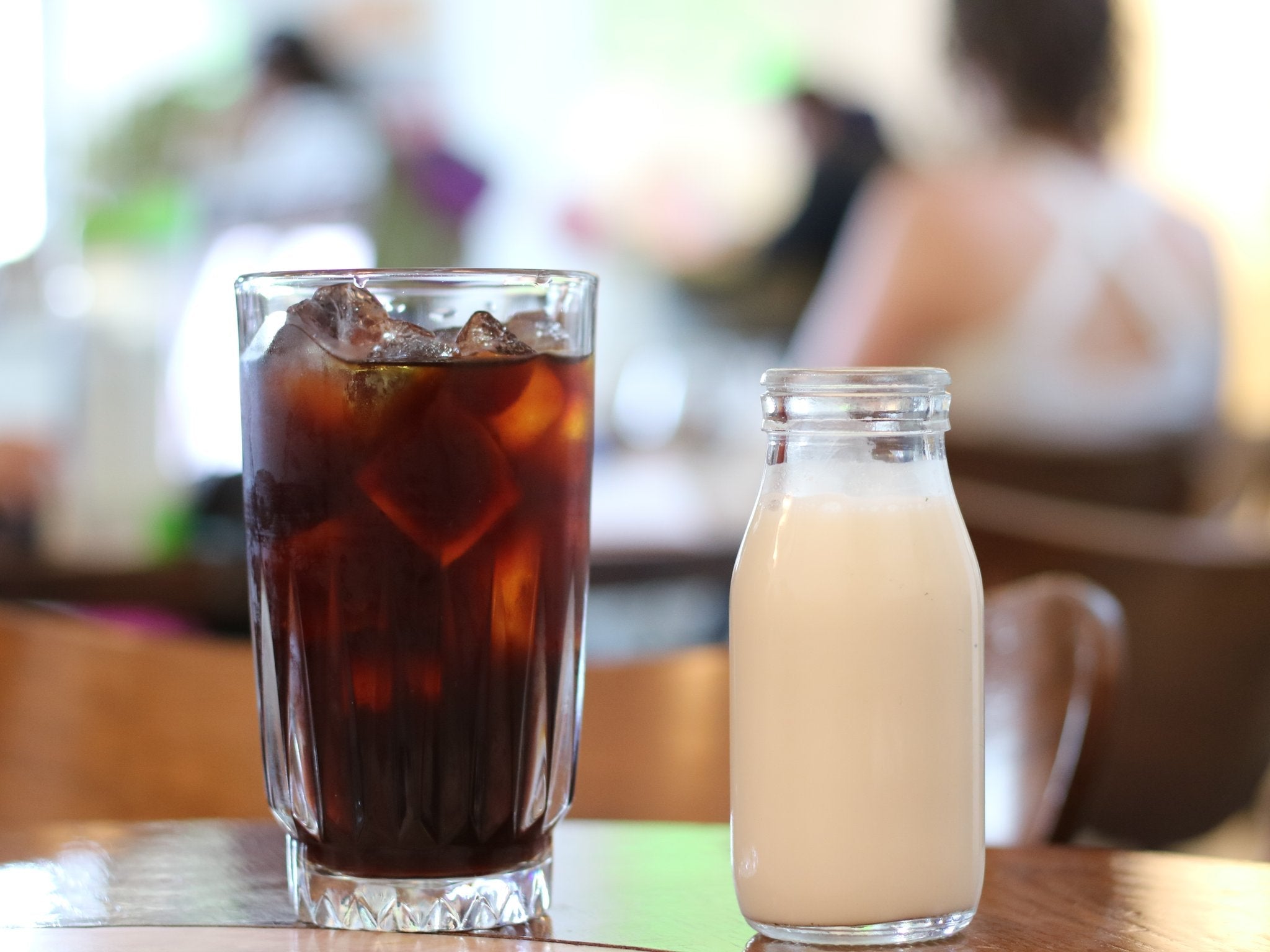 A tall glass of iced coffee and a bottle of light-colored JOI Vanilla Maple Creamer on a wooden table.