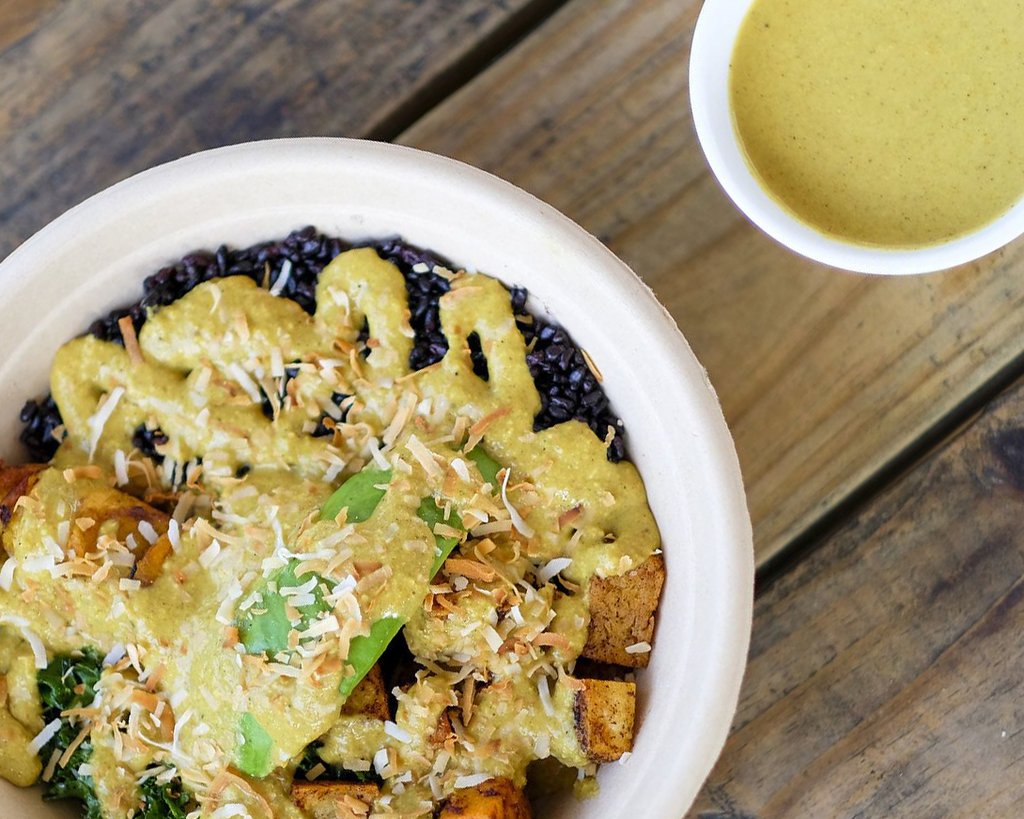 A DELLA Bowls yellow sauce-covered veggie bowl with black rice, roasted vegetables, and shredded coconut, with a side of extra sauce.