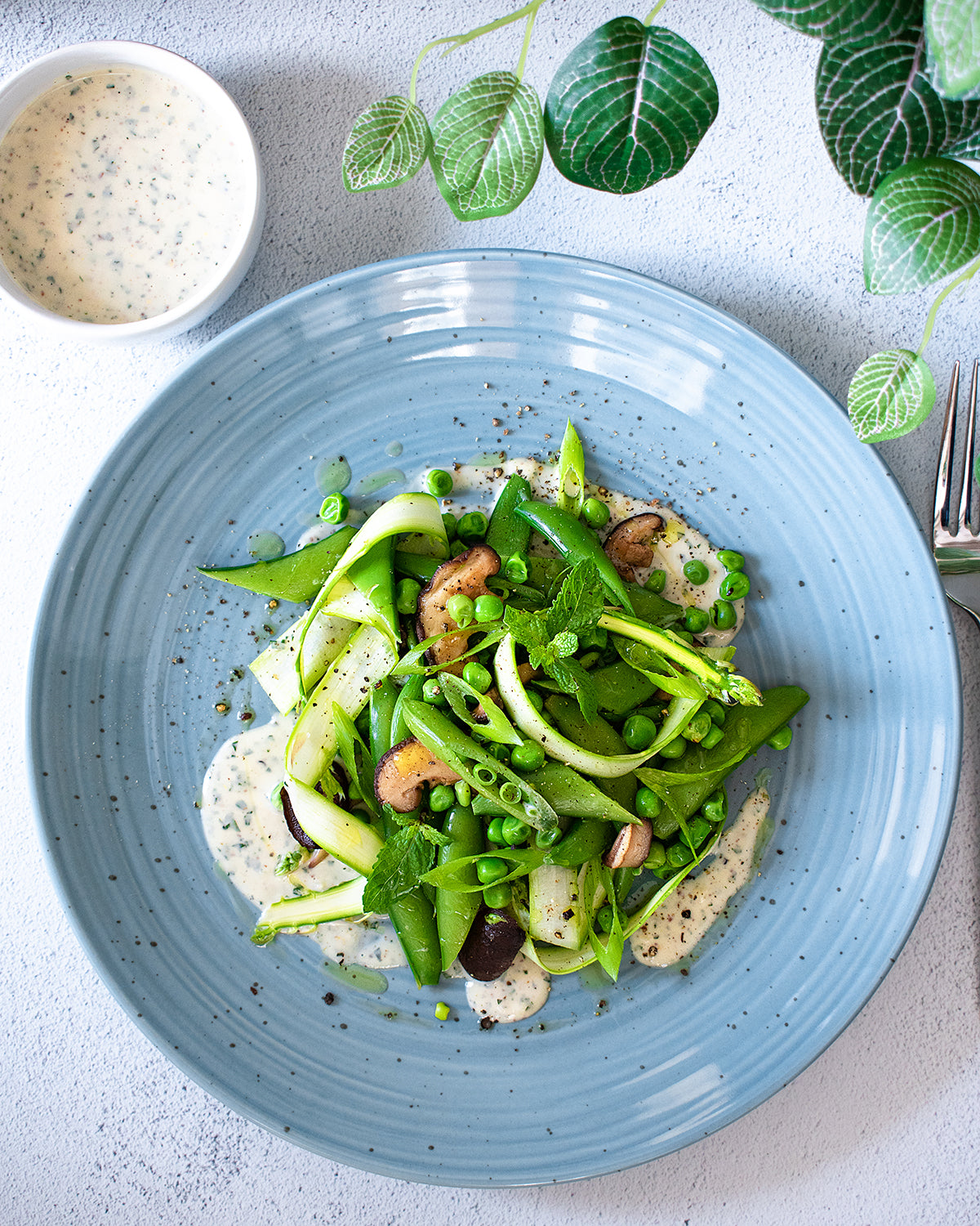 A fresh Spring Peas and Mushrooms with Mint Dressing dish featuring asparagus, sugar snap peas, and garden peas on a blue plate, with a side of JOI plant base dressing.