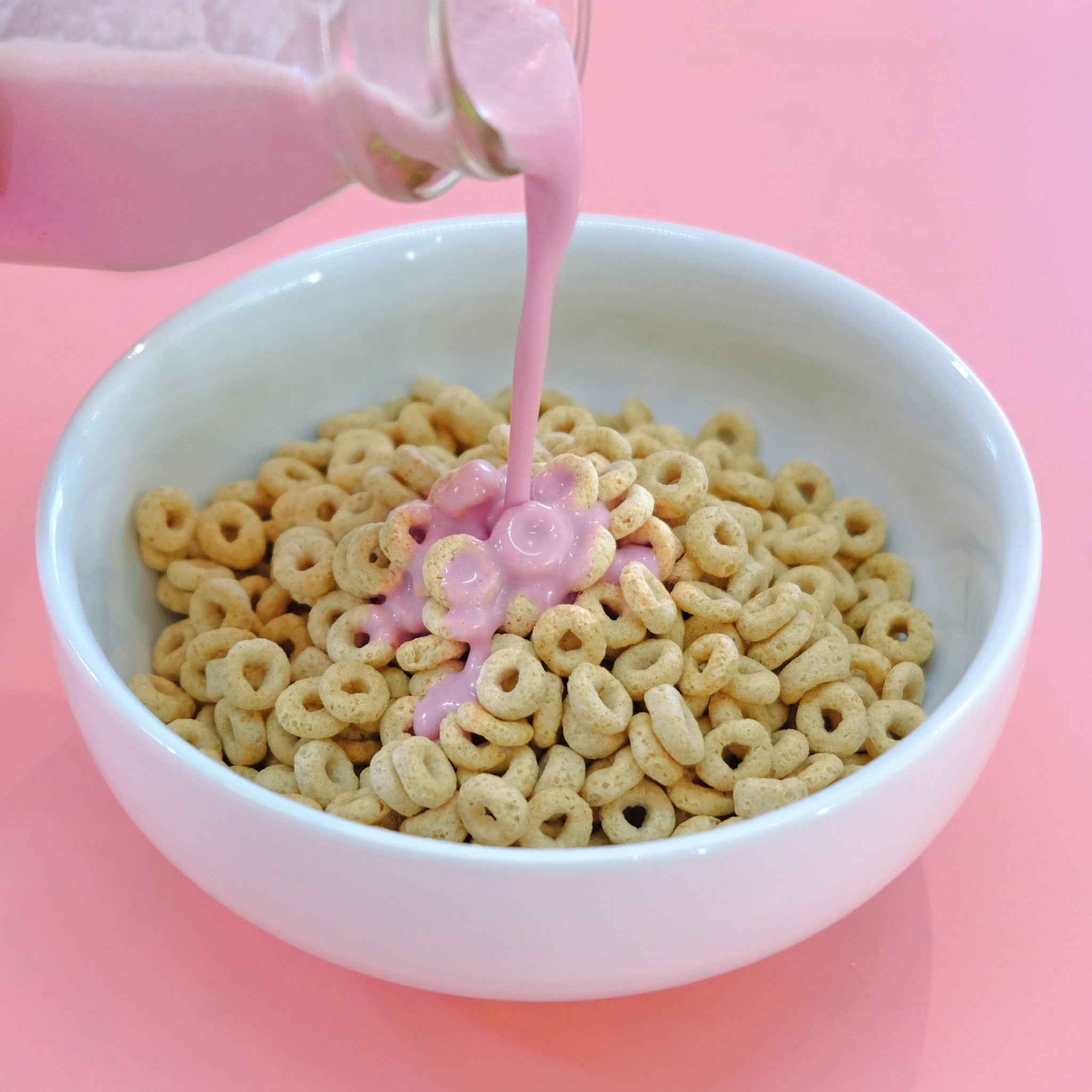 Pink JOI strawberry milk is poured into a white bowl of cereal loops on a pink background.