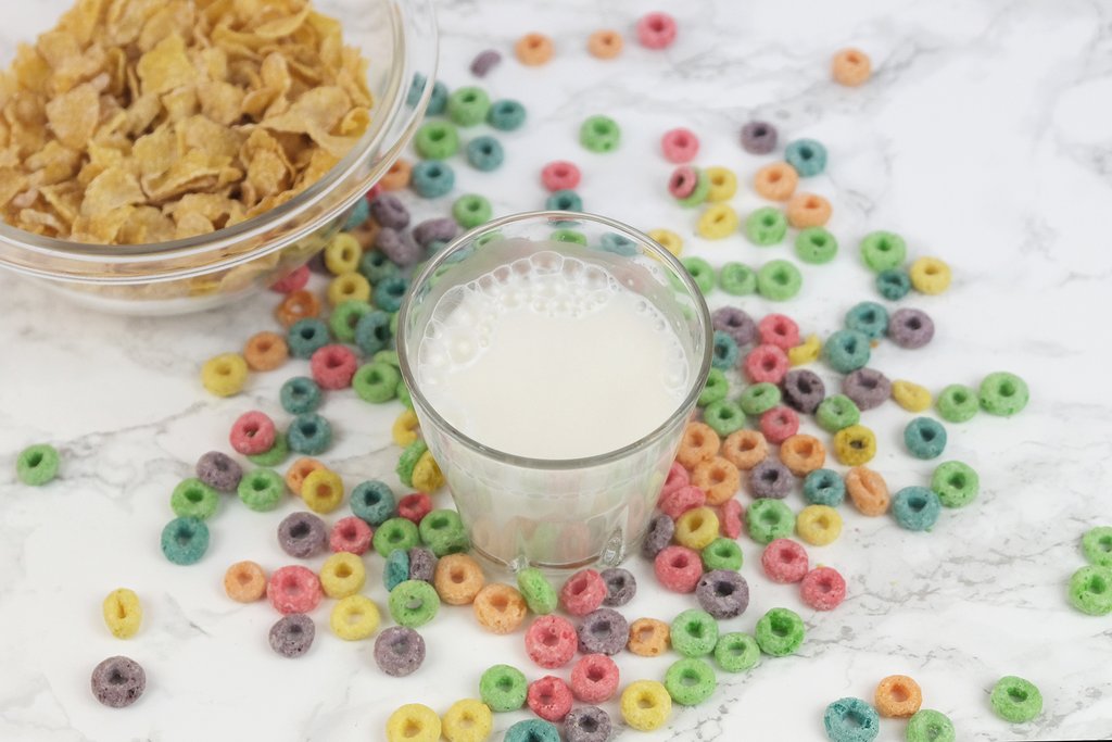 A glass of JOI plant-based milk sits among scattered colourful cereal rings, with a bowl of cornflakes nearby.