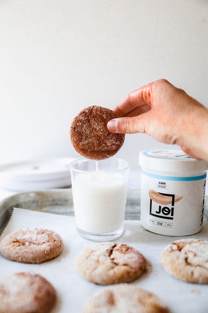 A hand dips an almond molasses crinkle cookie into a glass of milk next to a JOI almond nutbase container.