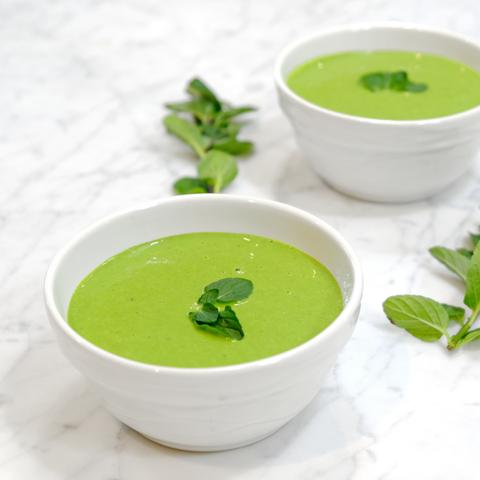 Two white bowls of JOI dairy-free creamy pea soup with mint on a white marble surface with mint sprigs.