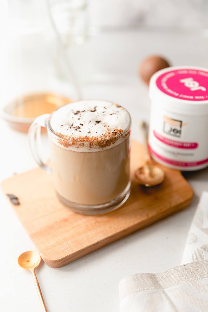 A JOI dairy-free Irish coffee cocktail, topped with foam and chocolate shavings, sits on a wooden board next to a JOI creamer tub.