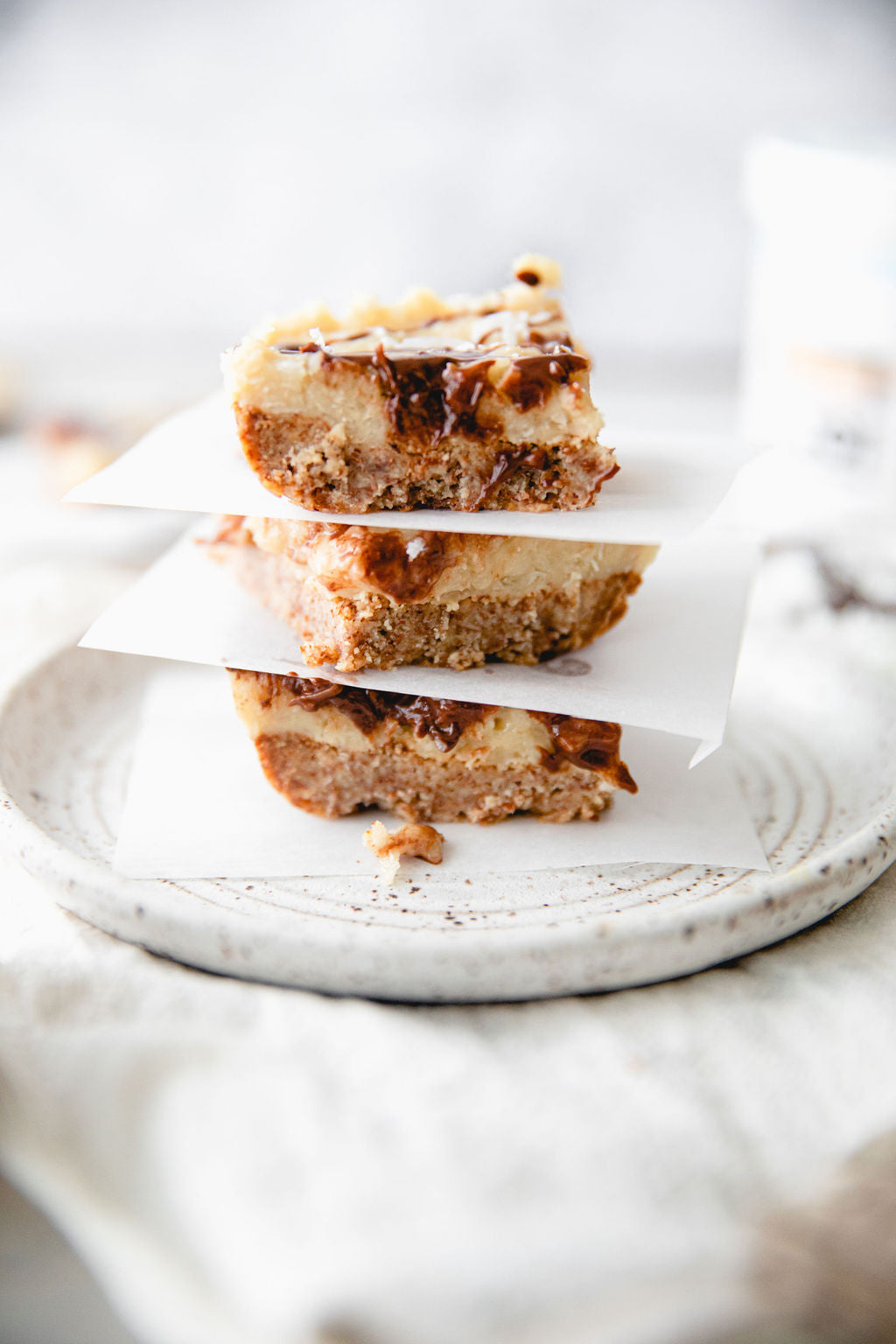 Three vegan Samoa bars, layered with a light base, white cream, and a rich caramel swirl, stacked on a white plate.