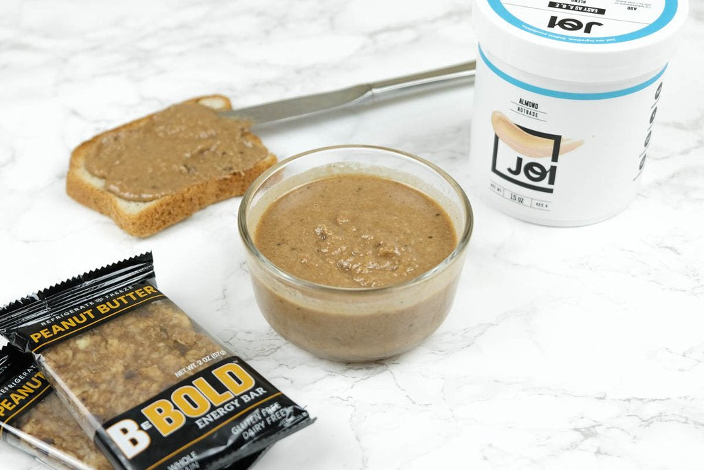 A bowl of JOI almond milk base energy butter with a slice of peanut butter toast and B-BOLD peanut butter energy bar.