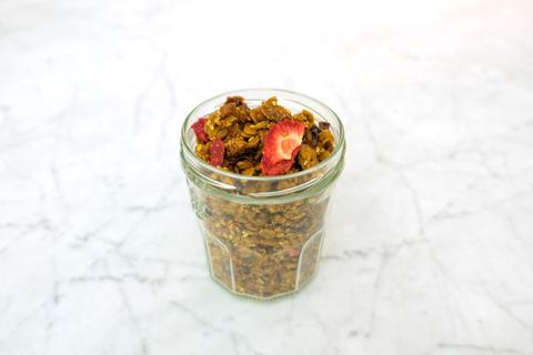 A glass jar filled with Joy The Baker's cashew-turmeric granola, topped with dried strawberries, on a white marble surface.