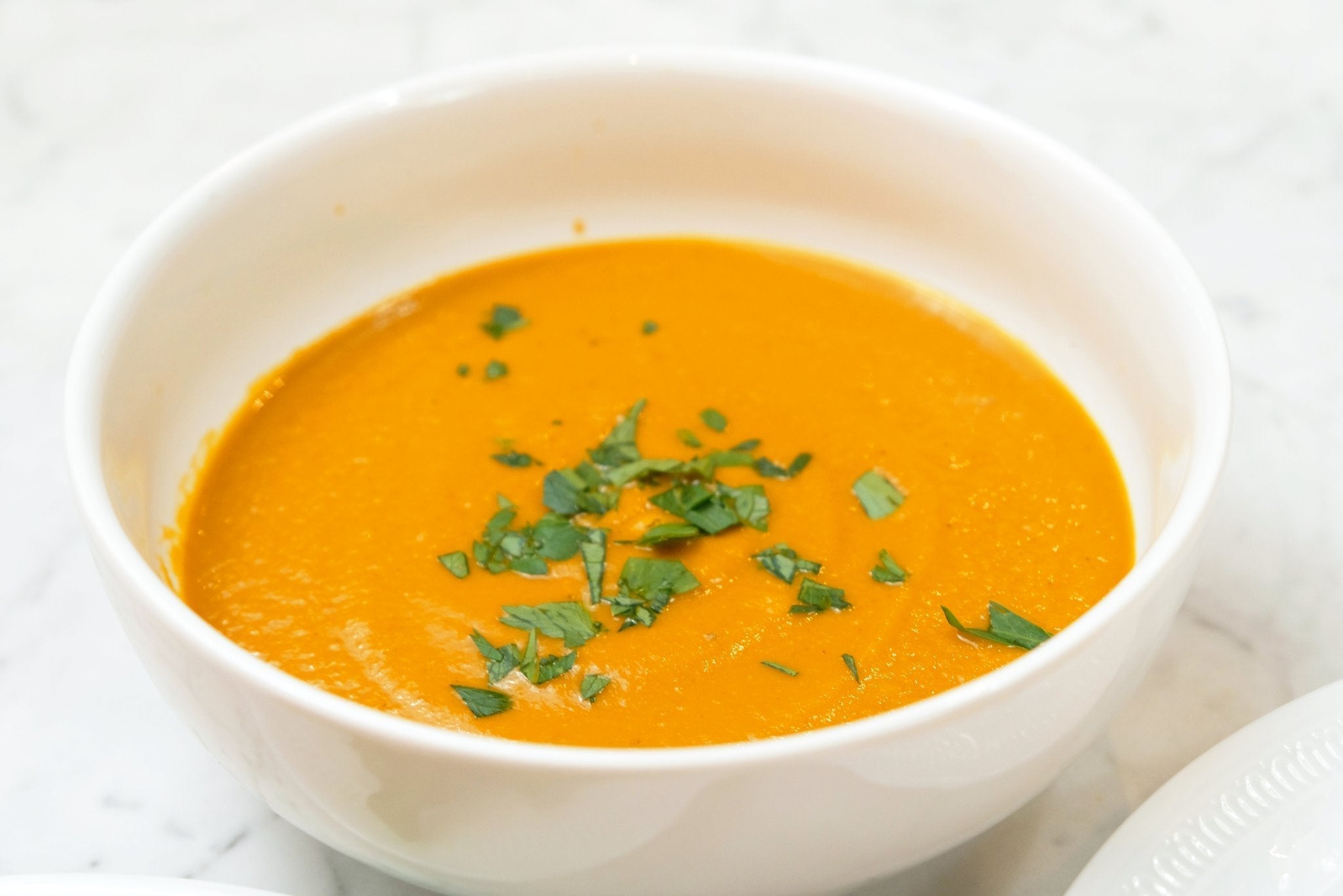 A white bowl filled with vibrant orange JOI Roasted Carrot and Turmeric soup, garnished with fresh green herbs.