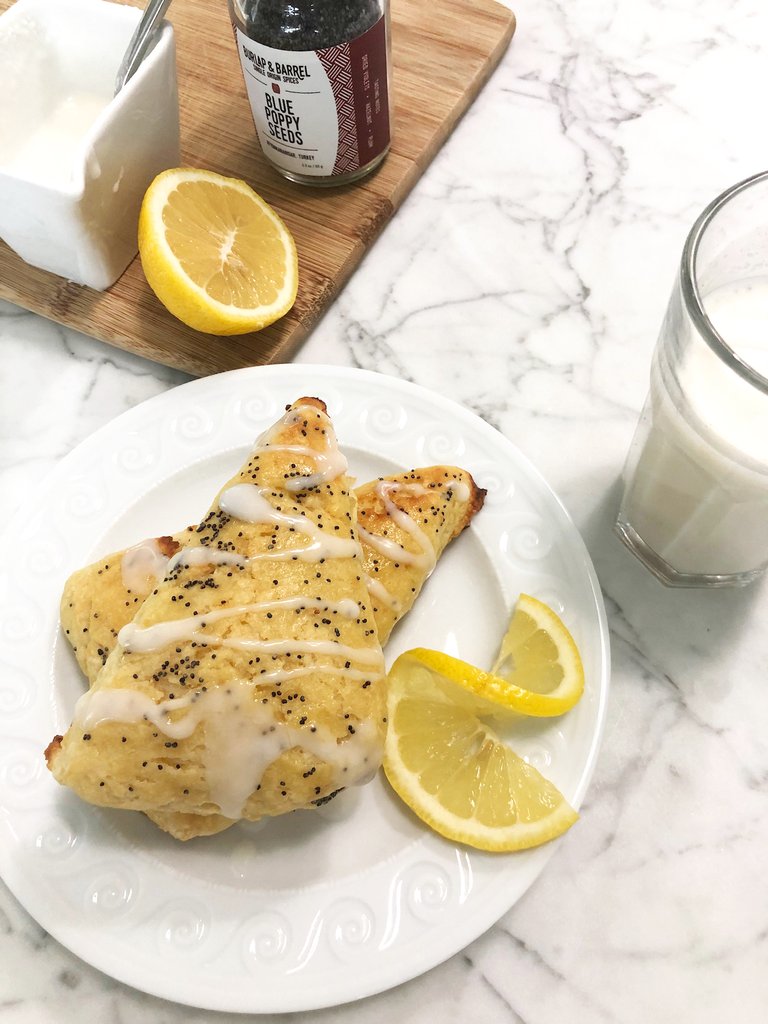Two glazed lemon poppy scones are drizzled with white icing and poppy seeds, served with lemon wedges and a glass of JOI plant milk.