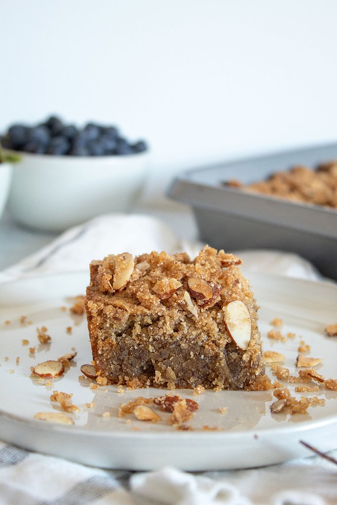 A delicious slice of JOI Vegan Toasted Almond Coffee Cake with crumb topping and sliced almonds, with blueberries in the background.