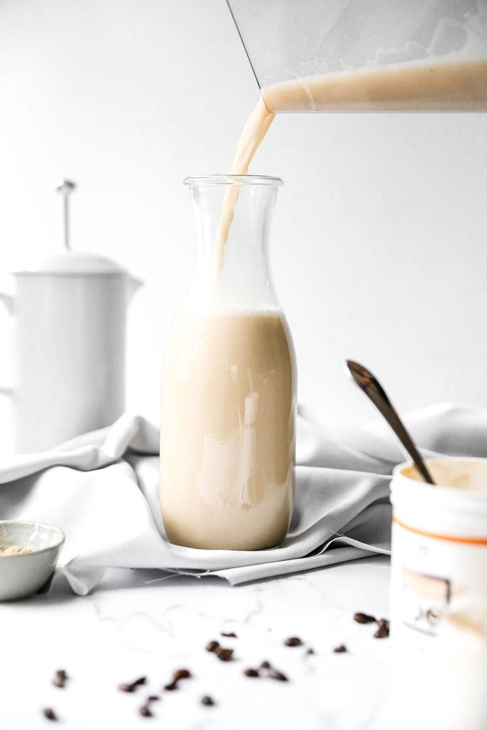 JOI Salted Caramel Creamer is poured from a blender into a glass bottle, with coffee beans scattered nearby.