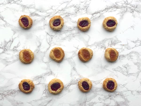 Twelve JOI Raspberry & Apricot Jam Almond Thumbprint Cookies with bright red and orange jam fillings are arranged on a white marble surface.