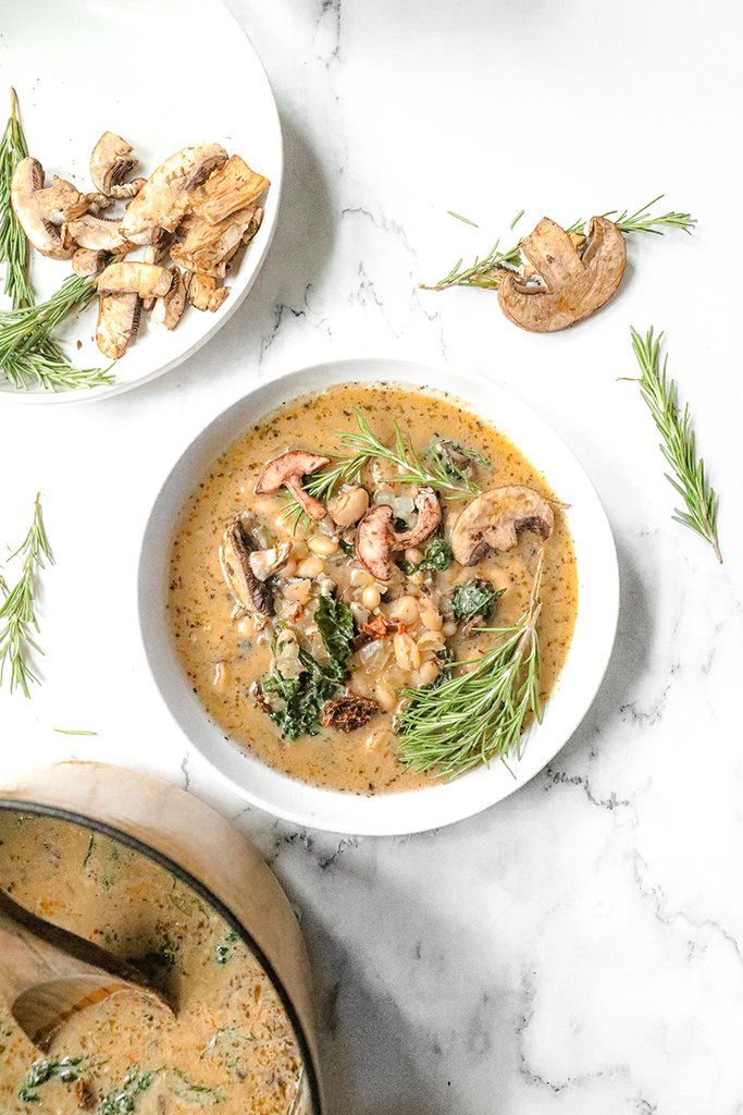 A bowl of creamy vegan white bean soup made with JOI, topped with roasted mushrooms and fresh rosemary, on a marble counter.