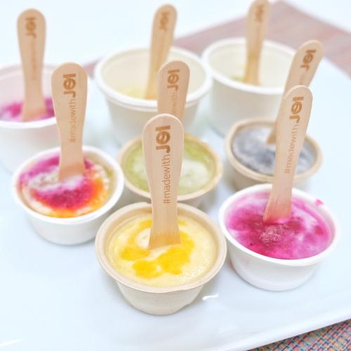 Nine small cups of colorful JOI plant-based coconut fruit popsicles on a white tray, each with a wooden spoon.