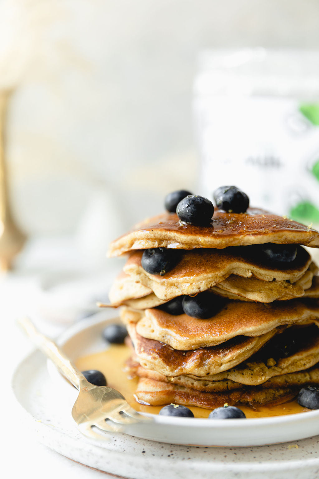 A stack of golden brown JOI Lemon Blueberry Pancakes drizzled with maple syrup and topped with fresh blueberries.