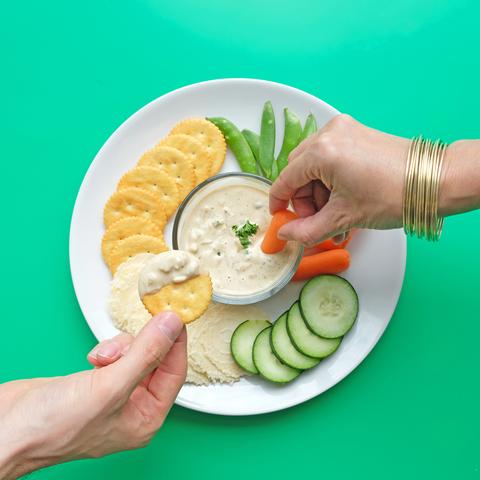 Hands dip crackers with JOI French Onion Dip and carrots into a bowl of creamy dip with fresh garnish.