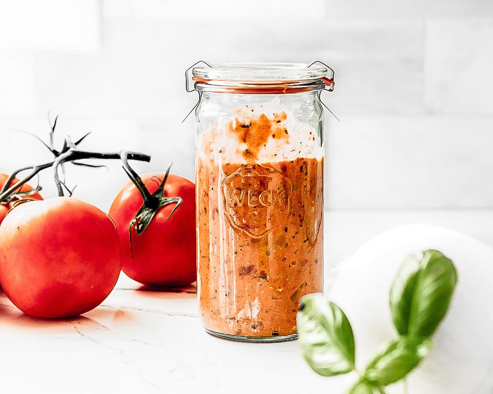 A tall glass Weck jar of creamy, reddish-orange JOI cashew-based skinny vodka sauce, with fresh tomatoes and basil.