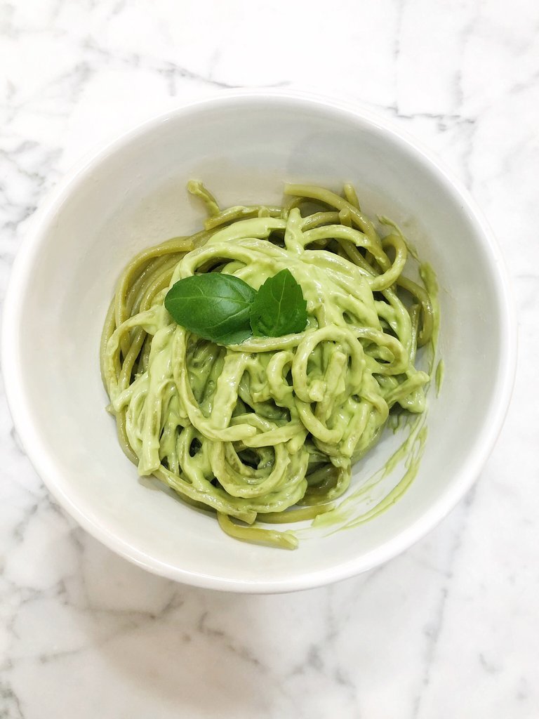 A white bowl on a marble surface holds green pasta coated in a creamy JOI avocado sauce and garnished with basil.