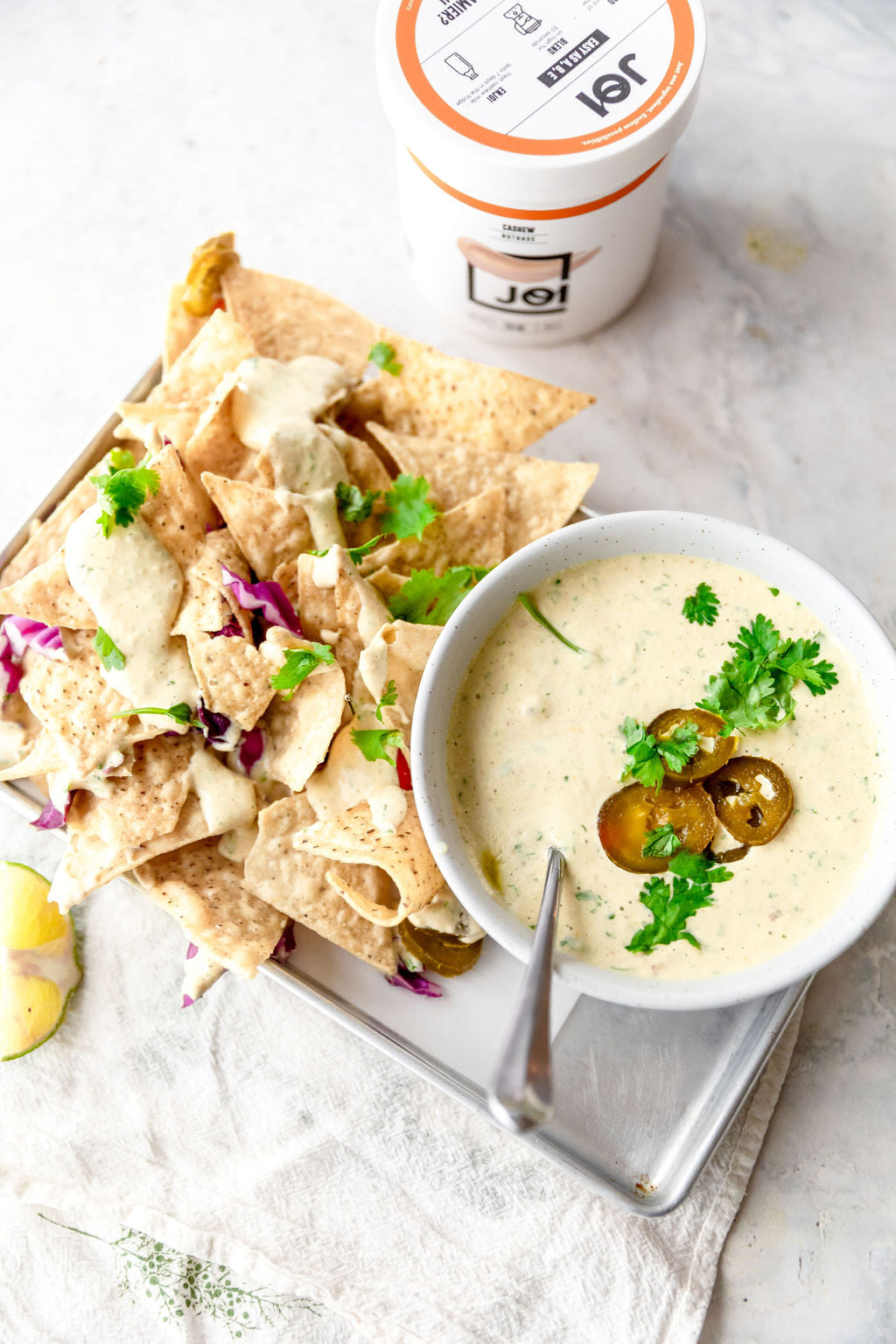 A bowl of JOI cashew plant-based white queso with pickled jalapeños, cilantro, and nachos on a baking sheet.