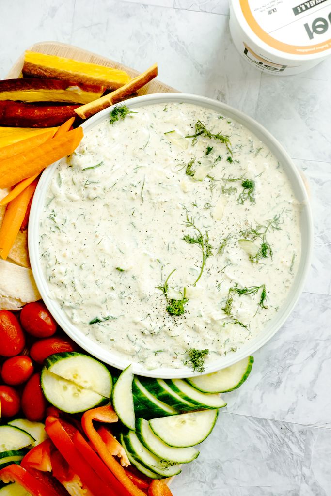 A creamy vegan, dairy-free, and gluten-free Greek Tzatziki Dip from JOI, garnished with dill and black pepper, surrounded by colorful fresh crudités and pita bread, with a JOI container visible in the background.