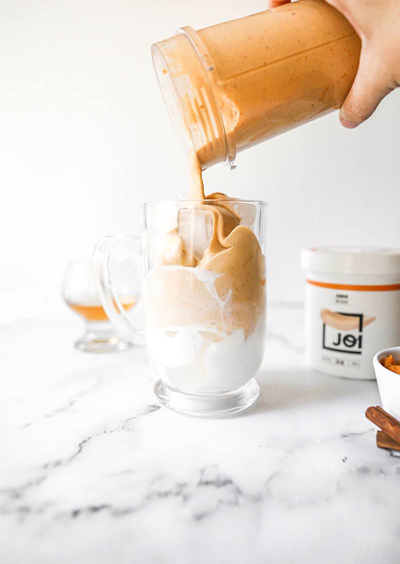 A hand pours creamy, orange JOI Pumpkin White Russian into a glass mug layered with white cream.