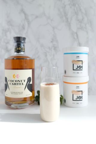 A bottle of Coconut Cartel rum, a glass of coquito, and two tubs of JOI plant base on a white marble surface.