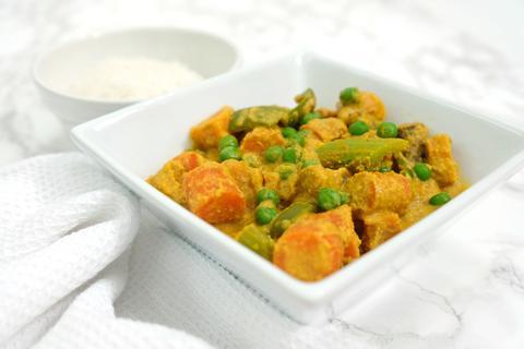 A white rectangular dish of vegan korma with sweet potatoes, green peas, and asparagus, served with rice.