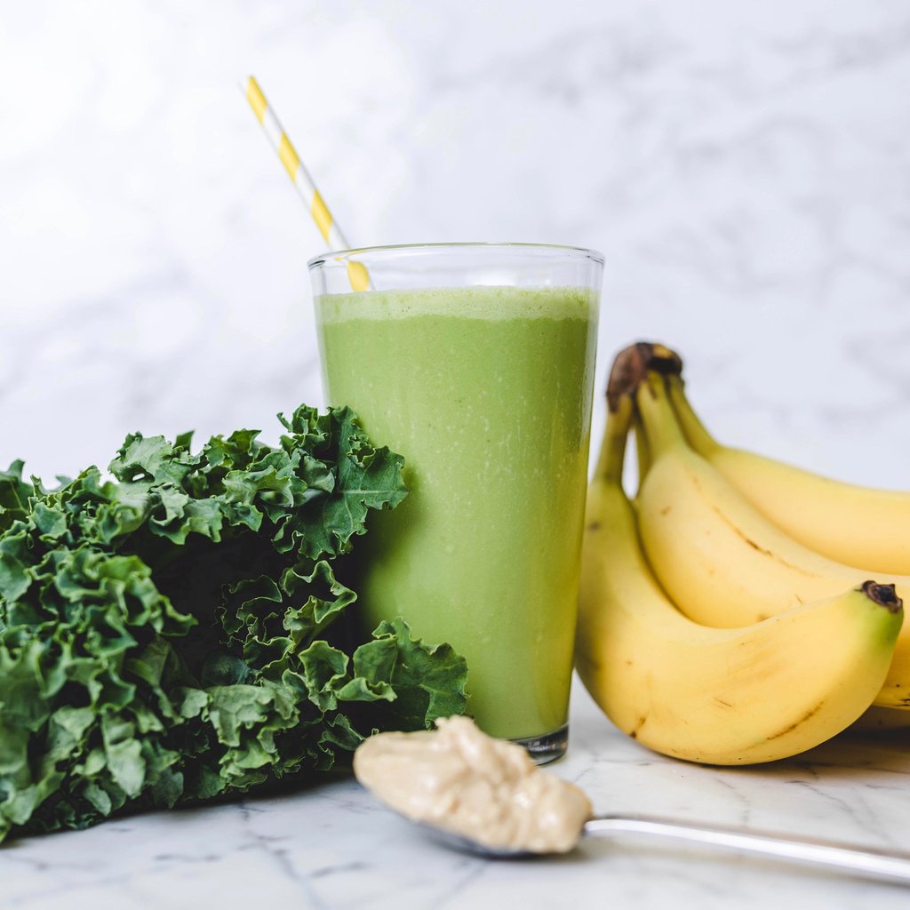 A vibrant green JOI smoothie with a yellow striped straw, fresh kale, bananas, and a spoonful of peanut butter.