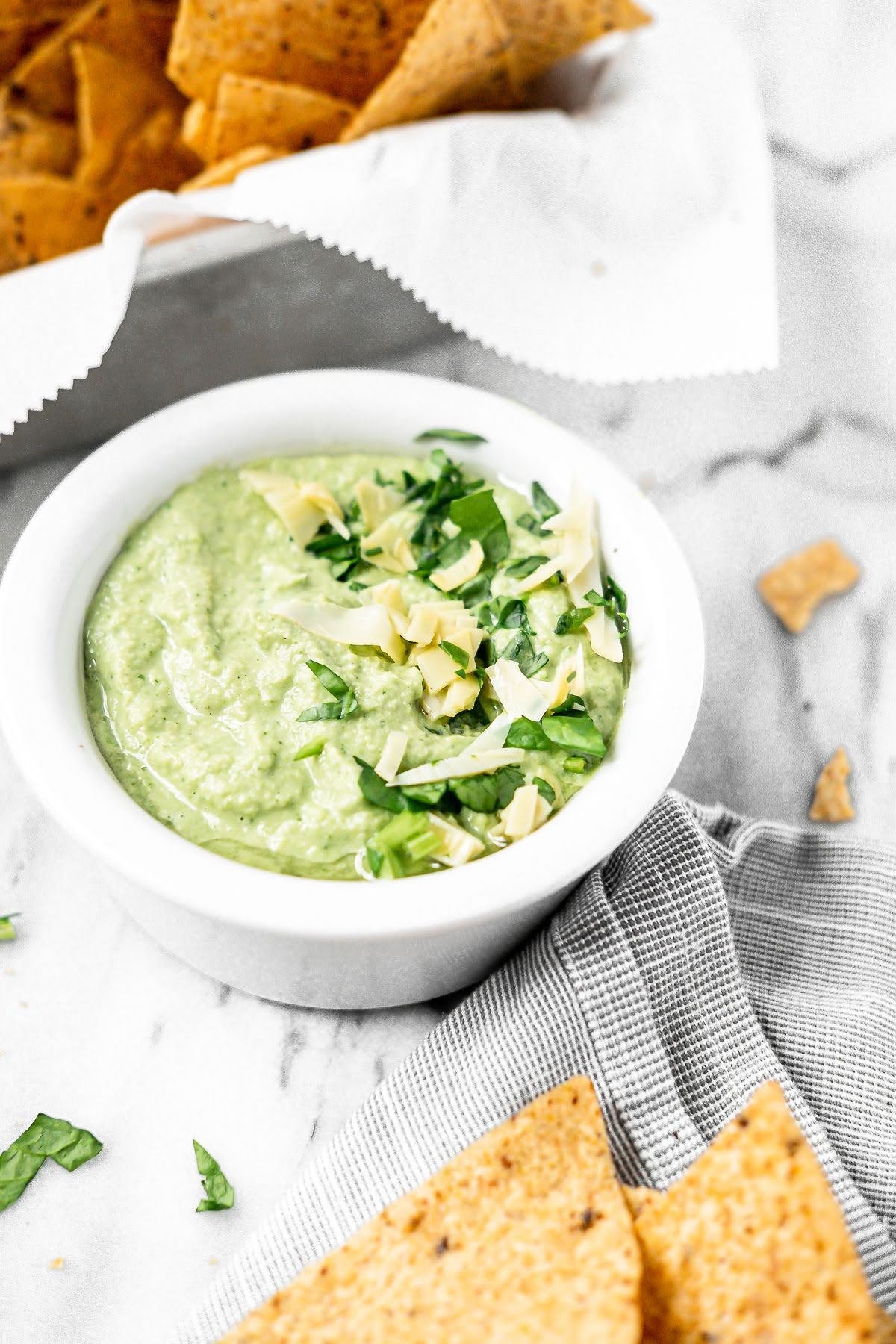 A white bowl of creamy, dairy-free spinach artichoke dip topped with herbs and cheese, next to tortilla chips.