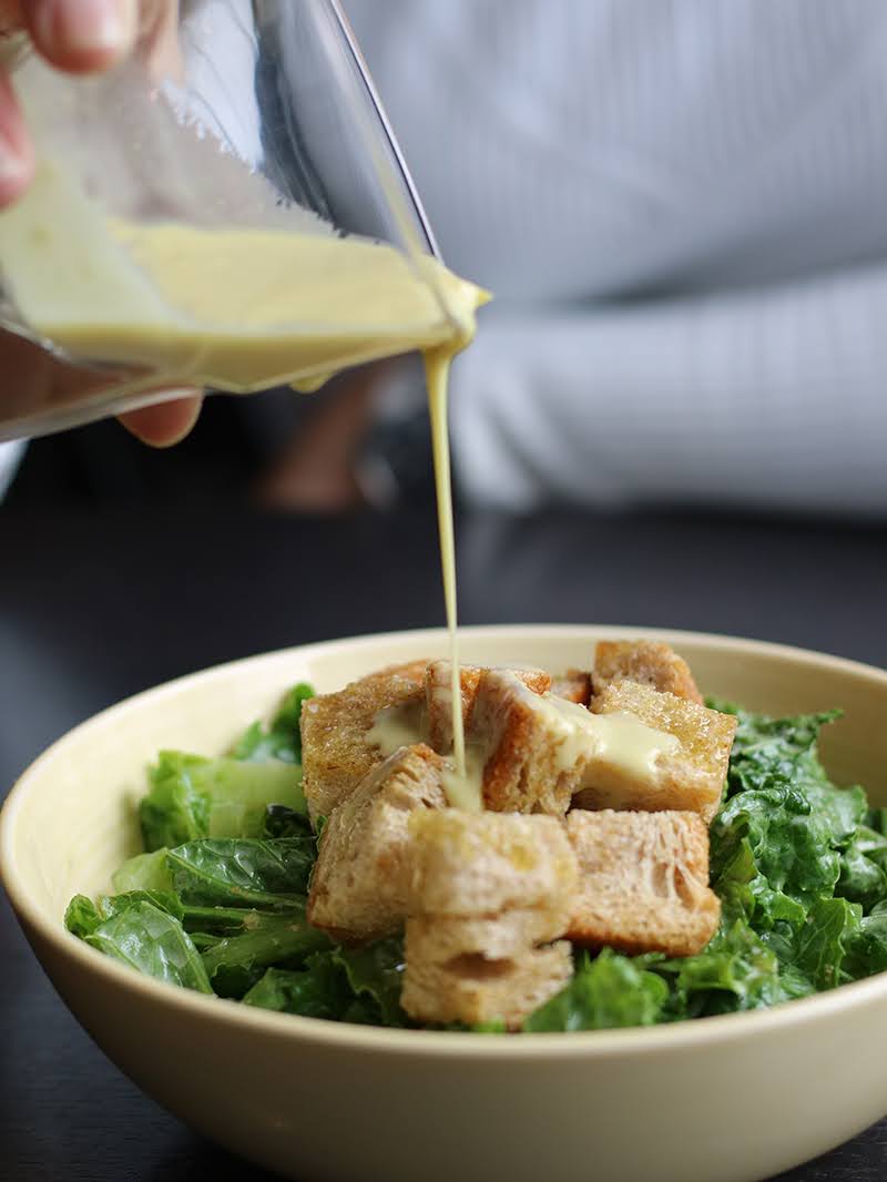 A rich, creamy JOI vegan Caesar dressing being poured from a glass onto a bowl of green lettuce and croutons.