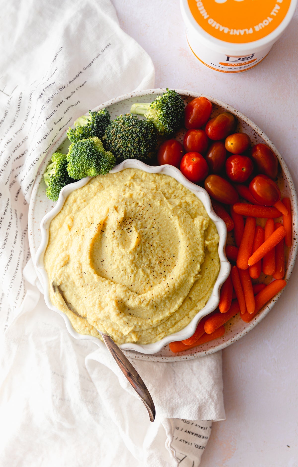 A bowl of dairy-free, vegan cauliflower dip surrounded by broccoli, cherry tomatoes, and baby carrots, with JOI plant base nearby.