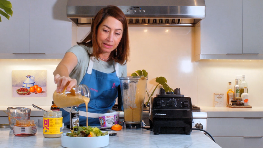 A woman pours creamy JOI Kimchi Dressing from a jar onto a salad, with a Vitamix blender nearby.