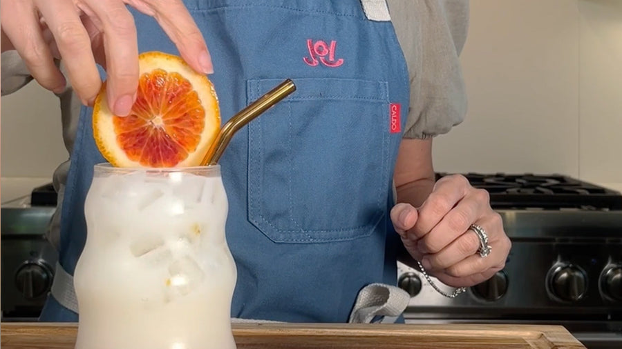 A hand garnishes a glass of JOI oat milk dirty soda with a blood orange slice and a metal straw.