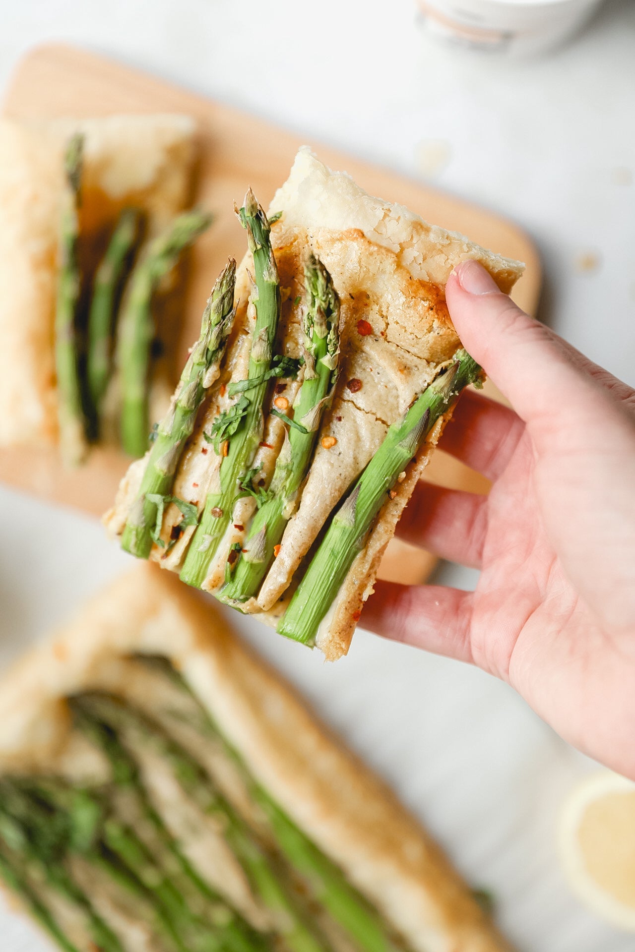 A hand holds a slice of JOI Asparagus and Cashew Puff Pastry topped with green asparagus spears and red pepper flakes.