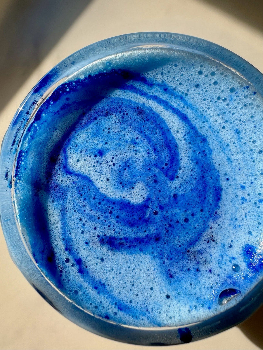 A vibrant blue JOI tropical smoothie, topped with white foam and a swirl of dark blue, in a clear glass.