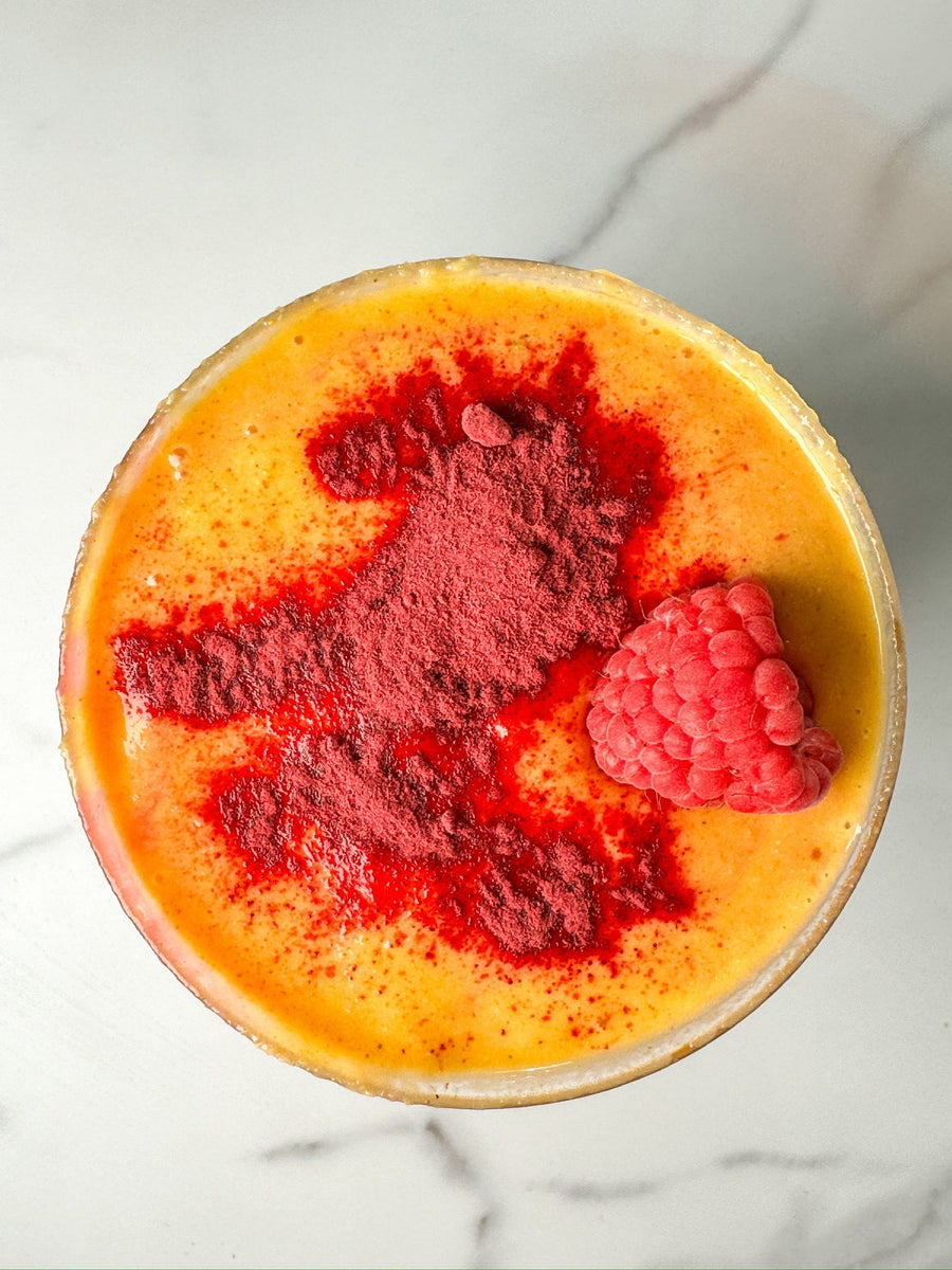 A JOI Golden Hour Peach Smoothie with a scoop of red powder and a fresh raspberry on a white marble surface.