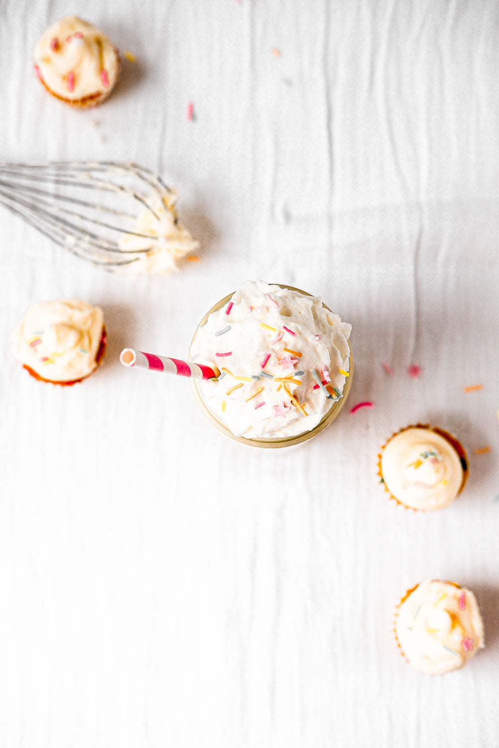 A creamy, dairy-free JOI Birthday Cake Shake with sprinkles and a striped straw, surrounded by mini cupcakes.