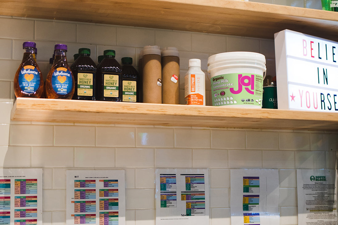 On a light wood shelf above a tiled wall, various organic honey bottles are next to a white bucket of JOI plant base.