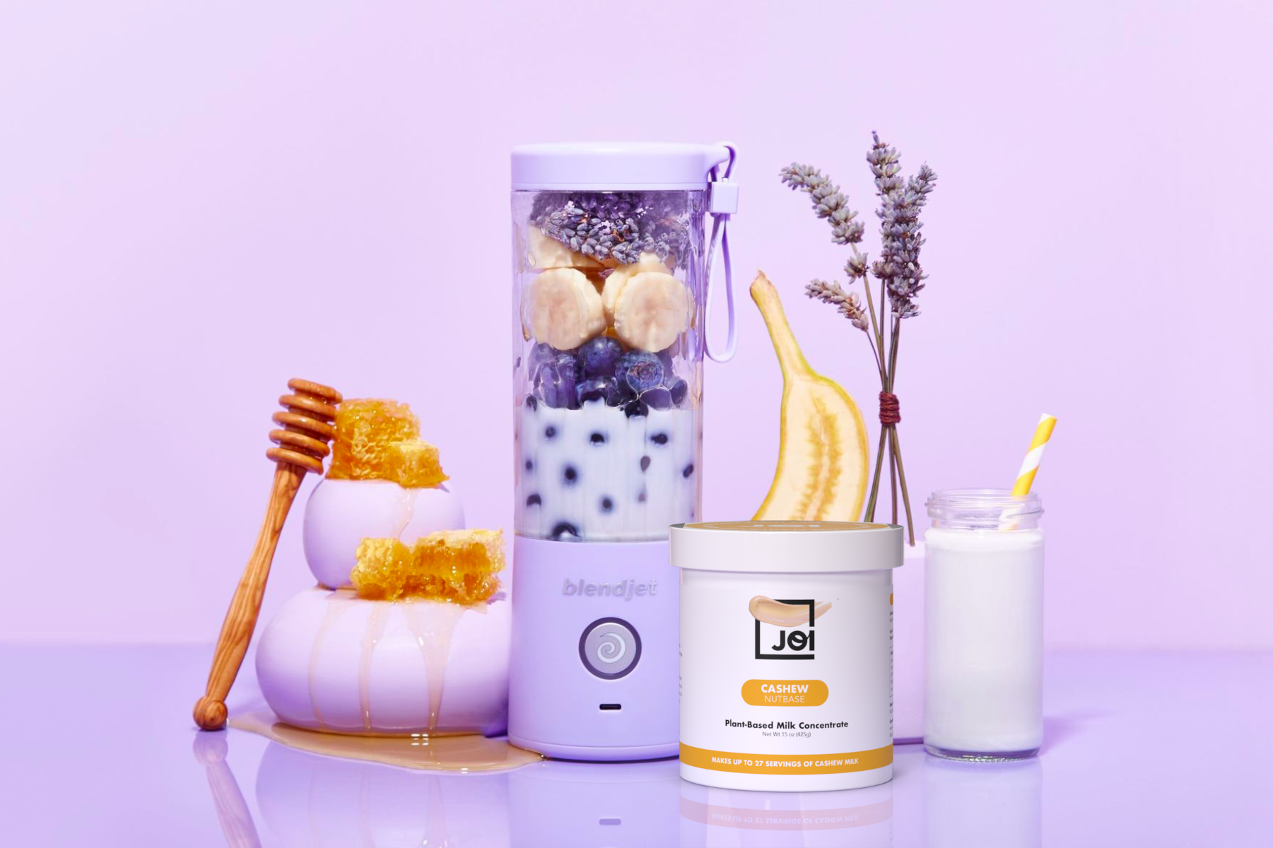 A lavender BlendJet 2 with a smoothie and a container of JOI Cashew Plant-Based Milk Concentrate are arranged with honey, a banana, and dried lavender.