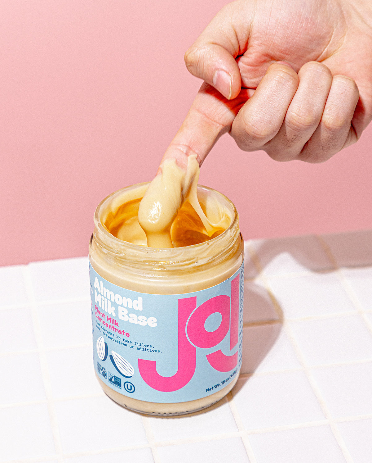 A finger drips creamy JOI Almond Milk Base from a jar with a blue label against a pink background.