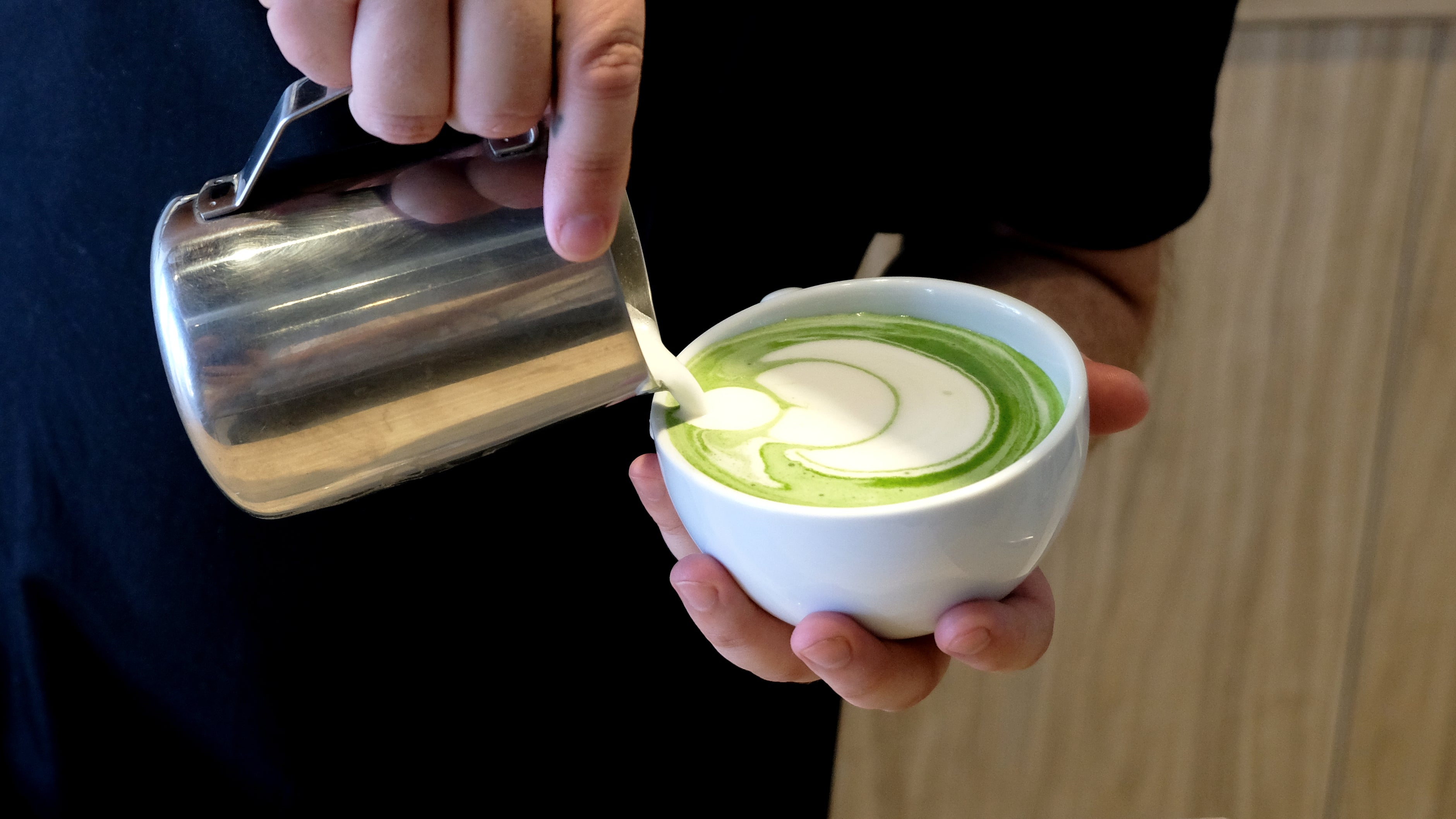hand pouring foamed milk out of a stainless container into a matcha in white ceramic