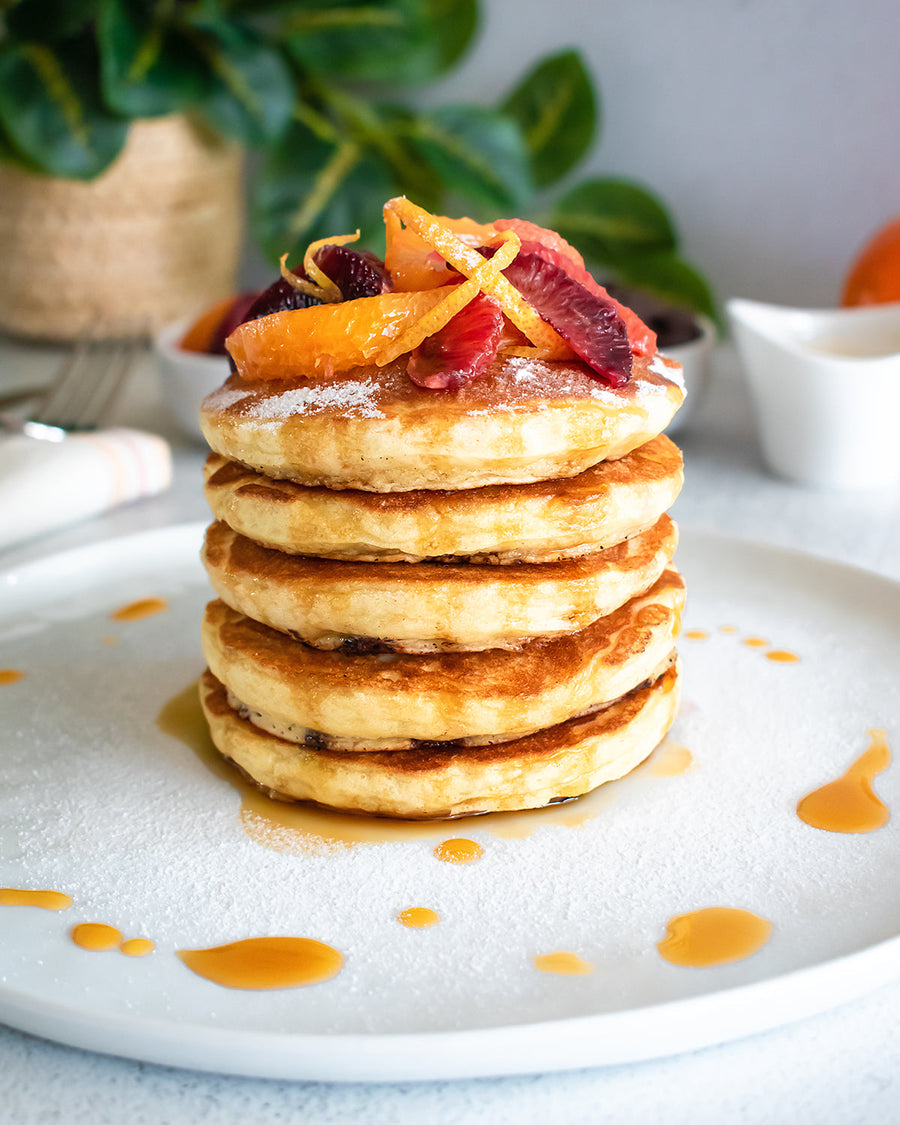 A stack of fluffy JOI vegan buttermilk pancakes with chocolate chips, topped with orange compote, powdered sugar, and syrup.