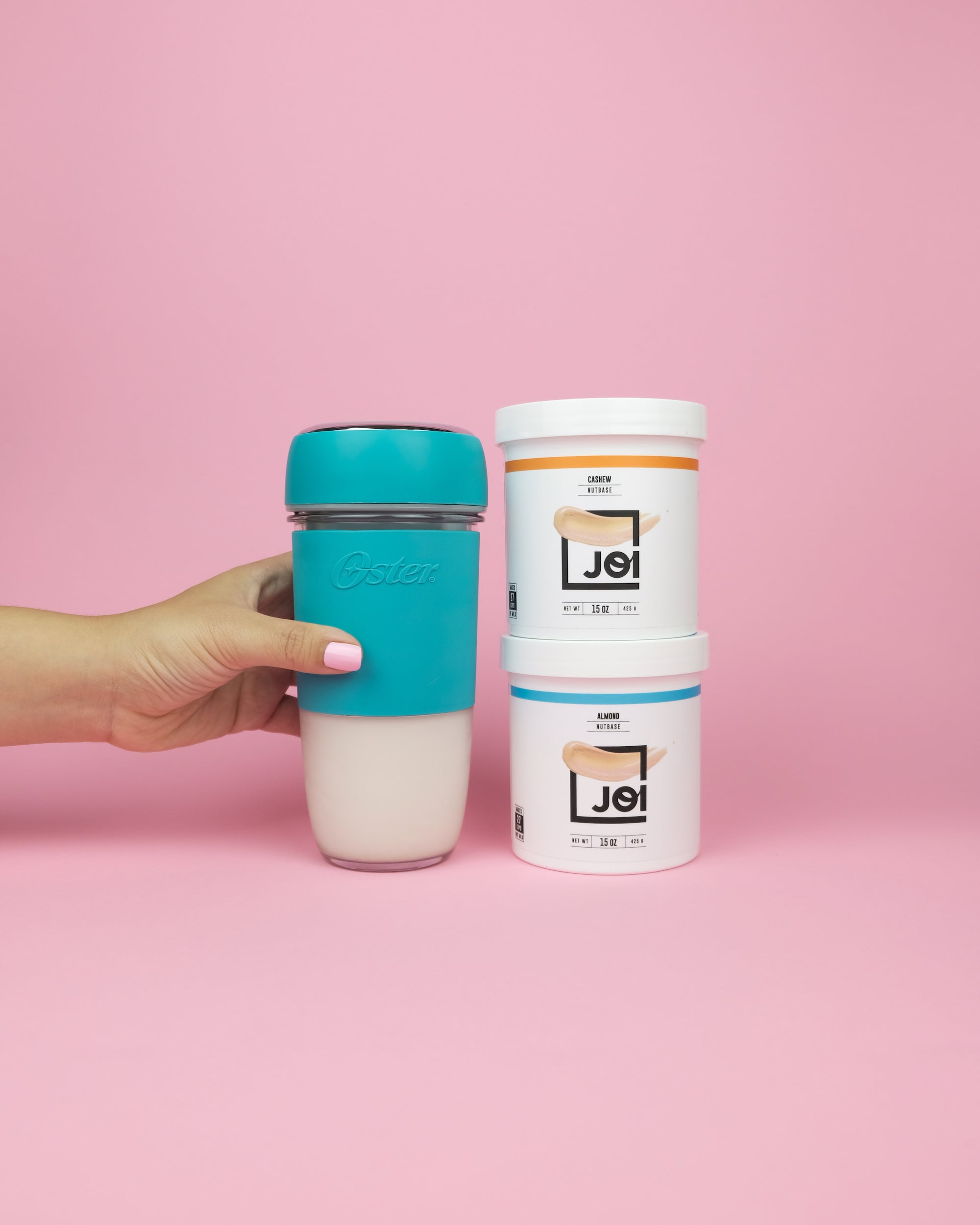A hand holds an Oster portable blender filled with JOI plant milk next to two tubs of JOI plant base concentrate.