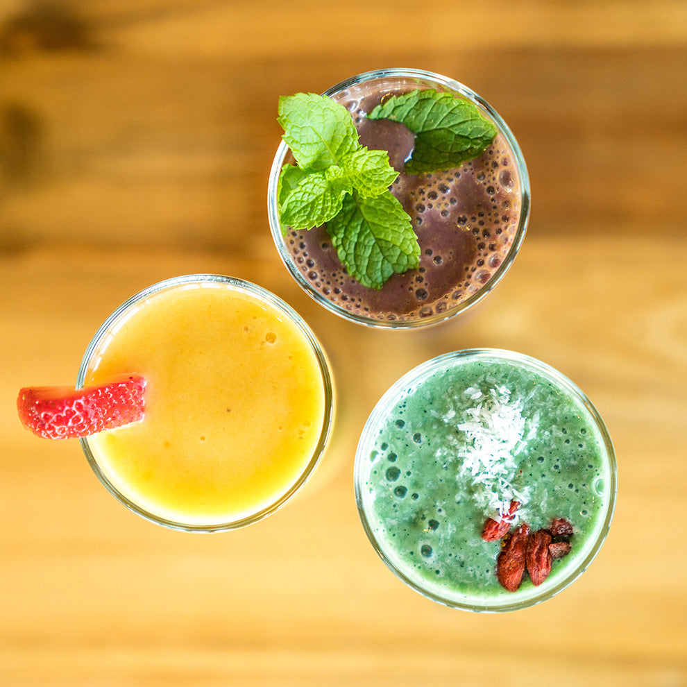 Three smoothies made with JOI plant bases: a purple-brown smoothie with mint, a yellow one with a strawberry, and a green one with coconut flakes and goji berries.