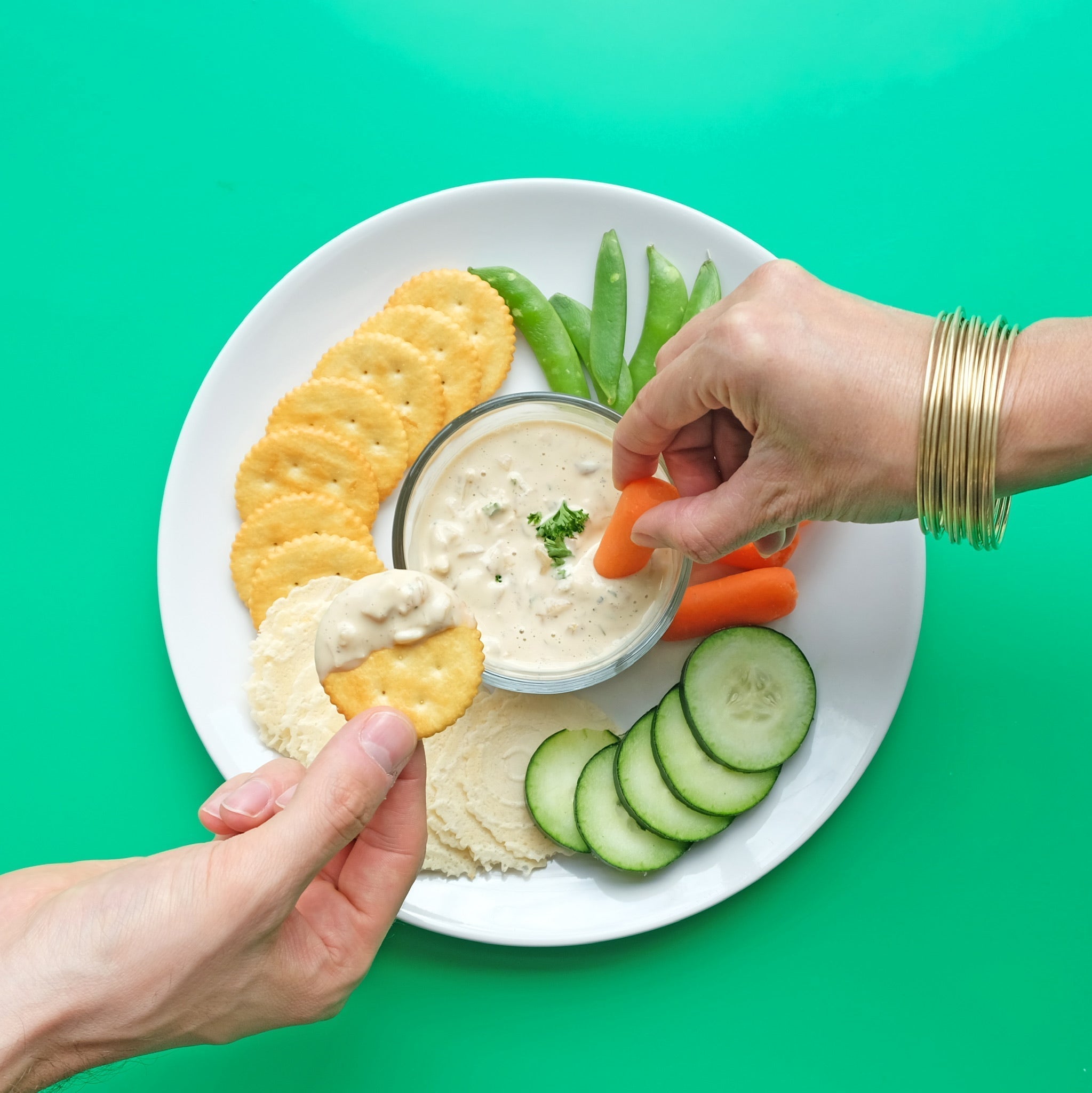 Two hands reach for a JOI plant-based dip, one with a baby carrot and another with a cracker, surrounded by vegetables.